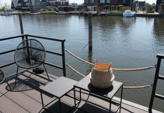 Uitzicht vanaf een woonboot bij Boat House Stavoren, Nederland, met stoelen, tafels en een mand aan het water.