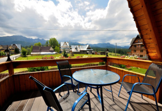 Uitzicht vanaf een chaletbalkon met blauwe stoelen en glazen tafel, omgeven door bergen en huizen in Tsjechië.