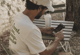 Mann mit Kappe sitzt draußen am Tisch, sieht auf sein Handy im Bell Tent bei Wecamp Jávea, Spanien.