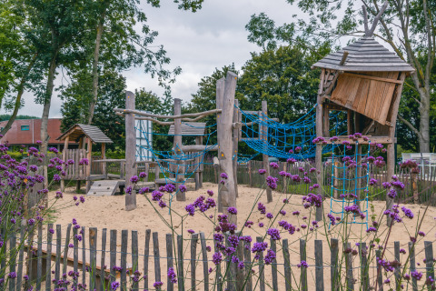 Speeltuin met zand, houten hutten en klimnetten, omringd door paarse bloemen bij een glampinglocatie.