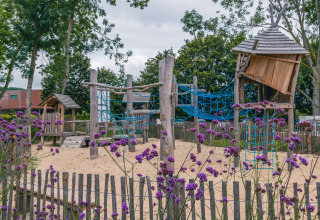 Speeltuin met zand, houten hutten en klimnetten, omringd door paarse bloemen bij een glampinglocatie.