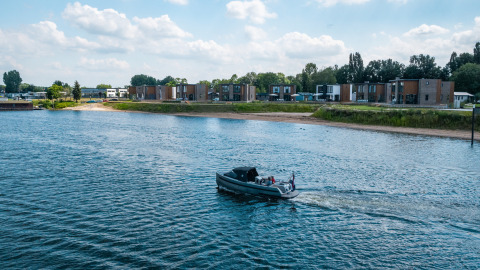 Sistemazioni glamping moderne sulla riva, con case in legno e una barca che naviga sul fiume tranquillo.