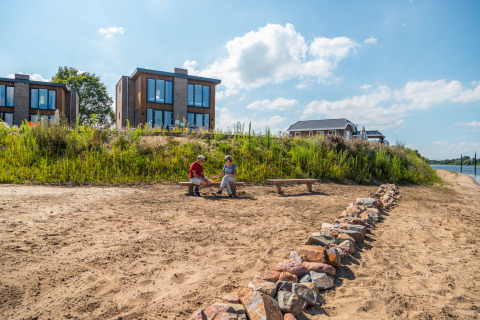 Deux personnes assises sur un banc sur une plage de sable devant des cabanes de glamping modernes au bord de l'eau.
