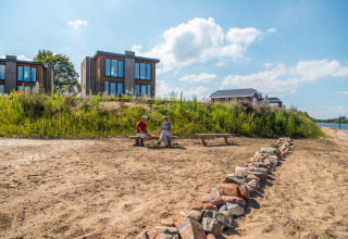 Due persone sedute su una panchina in una spiaggia sabbiosa davanti a lodge glamping moderni vicino all’acqua.