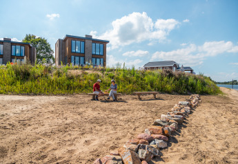 Zwei Menschen sitzen auf einer Bank am Sandstrand vor modernen Glamping-Häusern am Seeufer.