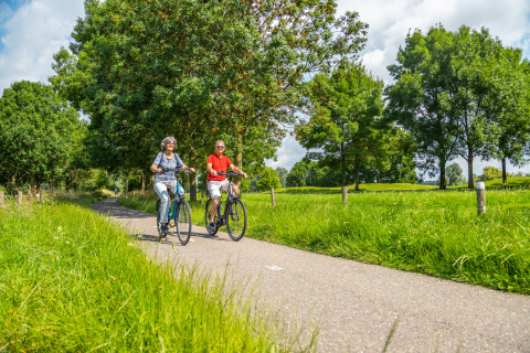 Een ouder echtpaar fietst samen over een groen pad bij een glamping accommodatie op een zonnige dag.