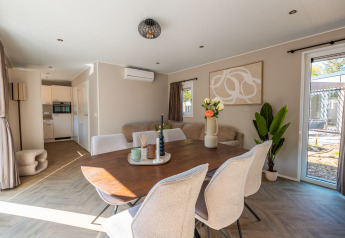 Moderne lodge met eetruimte, lichte stoelen, planten en ramen met zicht op een groene tuinomgeving.