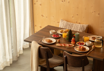 Desayuno en una cabaña con bollería, jugo de naranja, frutas y velas sobre una mesa de madera oscura.