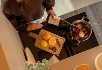 Une personne prépare du vin chaud sur la plaque, avec oranges, citron, épices et planche à découper à côté.