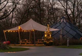 Scena all'aperto alla Unbrick One Lodge con tenda, luci, tavoli e sauna con piscina immersa nel bosco.