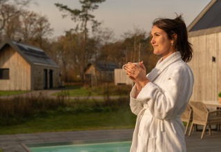 Mujer en bata disfruta de una bebida caliente junto a la piscina en Unbrick One lodge en De Weelderik, Países Bajos.