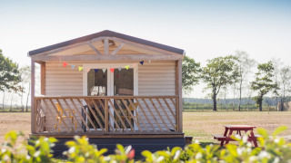 Beach Lodge con veranda in legno, festoni colorati e tavolo da picnic rosso in un paesaggio aperto.