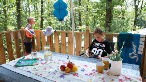 Deux garçons profitent de la terrasse d’une Lodge Tent Deluxe en Italie avec boissons, fruits et forêt.