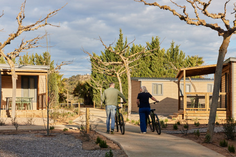 Twee personen met fietsen wandelen op het pad tussen houten huisjes op TAIGA Lake Caspe vakantiepark, Aragón, Spanje.