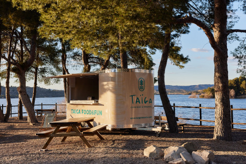 Foodtruck en picknicktafels aan een meer met bomen bij TAIGA Lake Caspe vakantiepark, Aragón, Spanje.