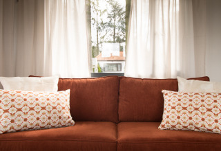 Knusse bruine sofa met sierkussens voor lichte gordijnen in Sun Lodge, Arden Parks Tulderheyde, België.