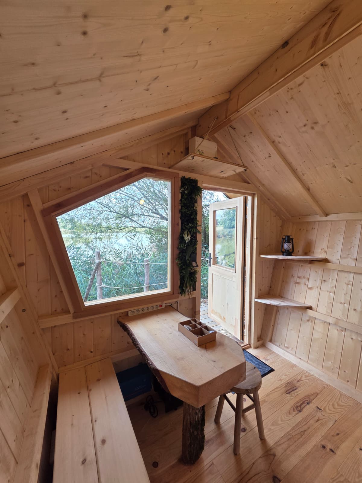 Knusse glamping hut met licht houten interieur, groot raam, rustieke tafel en zicht op de natuur buiten.