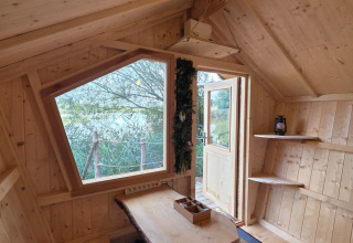 Knusse glamping accommodatie met licht houten interieur, groot raam, tafel en zicht op de natuur buiten.
