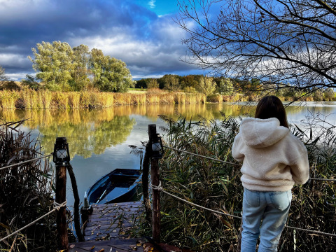 Persoon in witte trui aan een steiger, uitkijkend over een mooie vijver bij een glamping locatie.