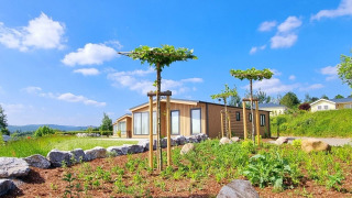 Chalet Enzian mit moderner Holzfassade, kleinen Bäumen und Steinen in sonniger Naturumgebung.