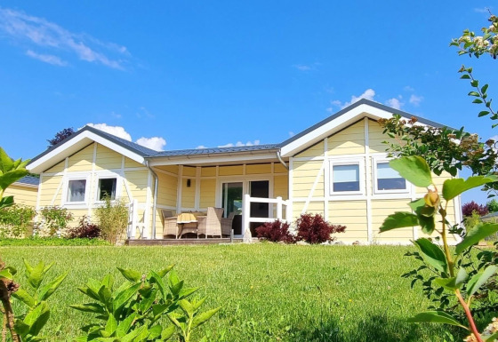Gele chaletwoning, Chalet Limonet, met overdekt terras in groene tuin onder een heldere blauwe lucht.