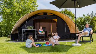 Familie genießt Zeit vor einem De Waard Tunneltent mit privatem Sanitär, Kinder spielen auf dem Rasen.