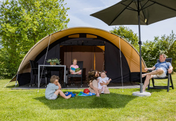 Famiglia rilassata davanti a una tenda safari De Waard con servizi igienici privati, bambini che giocano sul prato.