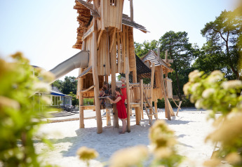 Parco giochi in legno in un glamping con bambini, adulto e natura verde attorno sotto il sole.