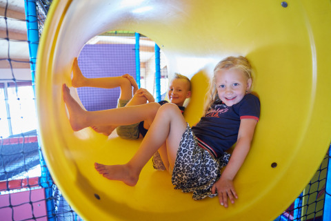 Kinderen spelen lachend in een gele glijbaan bij een indoor speelzone op een glampingverblijf.