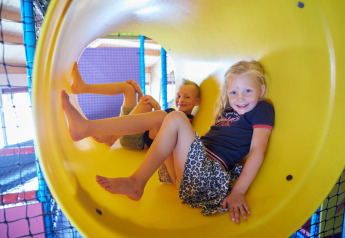 Niños jugando sonrientes dentro de un tobogán amarillo en un área de juegos de glamping.