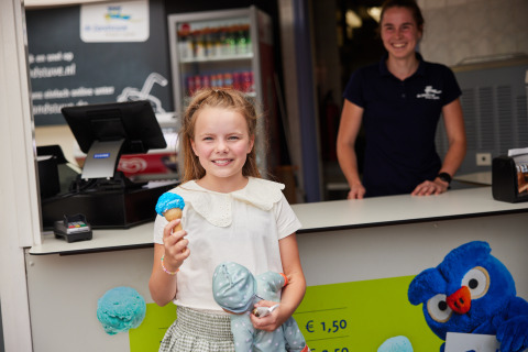 Fille avec une glace bleue en cornet et une peluche devant un comptoir, staff souriant sur un site de glamping.