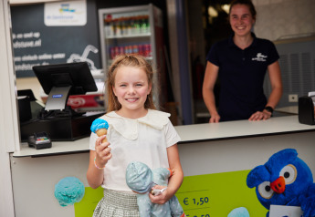 Bambina con gelato blu in cono e peluche davanti al banco gelati, staff sorride presso un glamping.