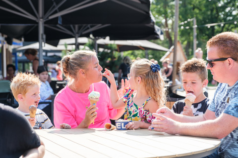 Een familie geniet samen van ijsjes aan een ronde buitentafel bij een safaritent op een zonnige dag.