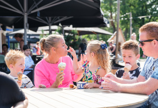 Een familie geniet samen van ijsjes aan een ronde buitentafel bij een safaritent op een zonnige dag.