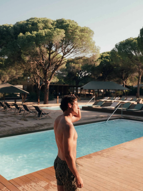 Giovane in costume si trova vicino alla piscina di Wecamp Cádiz, parco vacanze in Andalusia, Spagna, tra pini.