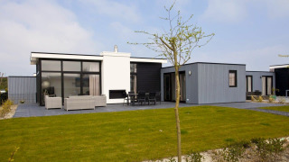 Lodge moderne L-Cube Hot tub avec grandes baies vitrées, terrasse meublée et pelouse verte, ciel bleu.