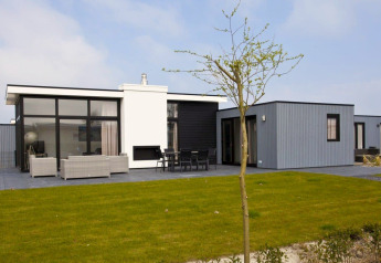 Lodge moderne L-Cube Hot tub avec grandes baies vitrées, terrasse meublée et pelouse verte, ciel bleu.