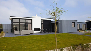 Moderne L-Cube Hot tub lodge met veel ramen, terras met zitbanken en een groene gazon in de zon.