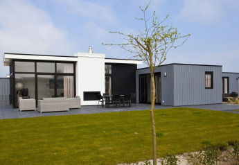 Moderne L-Cube Hot tub lodge met veel ramen, terras met zitbanken en een groene gazon in de zon.