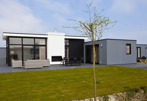 Moderne L-Cube Hot tub lodge met veel ramen, terras met zitbanken en een groene gazon in de zon.