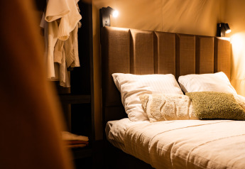 Vista interior de una tienda safari con cama cómoda, cabecero acolchado y luces de lectura cálidas.