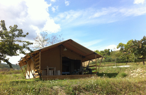 Safari tent with sanitary facilities in a grassy field, surrounded by trees and under a blue sky.
