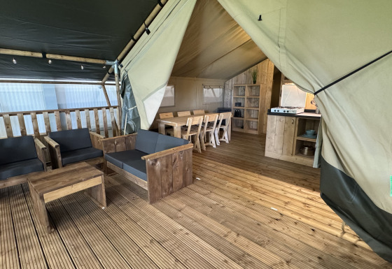 Interior of a safari tent with wooden deck, lounge area, dining table, and kitchenette with amenities.