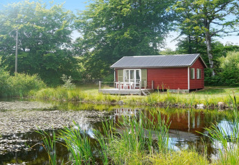 Rotes Ferienhaus am Teich mit Terrasse, umgeben von Bäumen im ruhigen Smedstorp, Schweden.