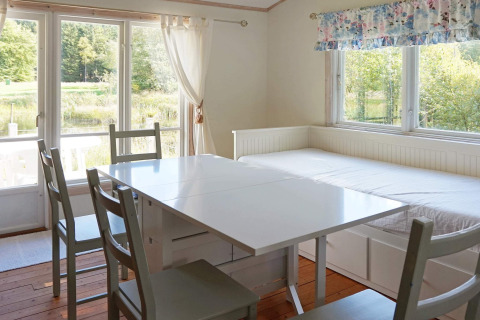Lyst spiseområde med store vinduer, udsigt til natur, spisebord, stole og sovesofa hos Smedstorp hytte i Sverige.