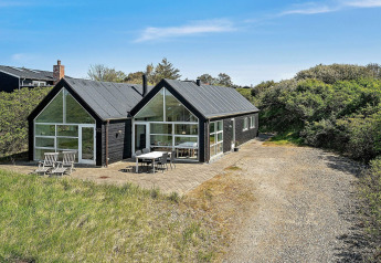 Modern zwart vakantiehuis in Blokhus, Denemarken, met grote ramen en terras met tuinmeubilair buiten.