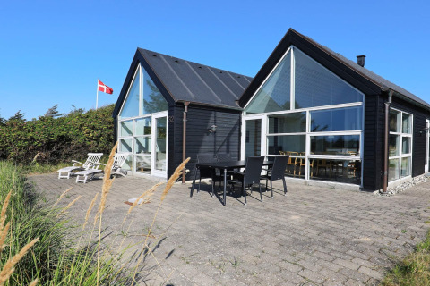 Moderne feriebolig i Blokhus, Danmark, med store vinduer, terrasse og dansk flag under blå himmel.