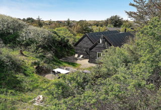 Sort træferiehus omgivet af grøn natur med udendørs terrasse og havemøbler i Blokhus, Danmark.