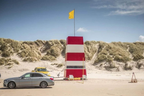 Foto van een rood-witte strandtoren met gele vlag, geparkeerde auto's en zandduinen op de achtergrond.