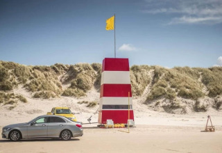 Foto van een rood-witte strandtoren met gele vlag, auto’s geparkeerd en zandduinen achteraan.
